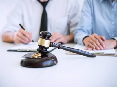 A gavel sits on a table with two gold wedding bands. Two people sign divorce papers in the background.
